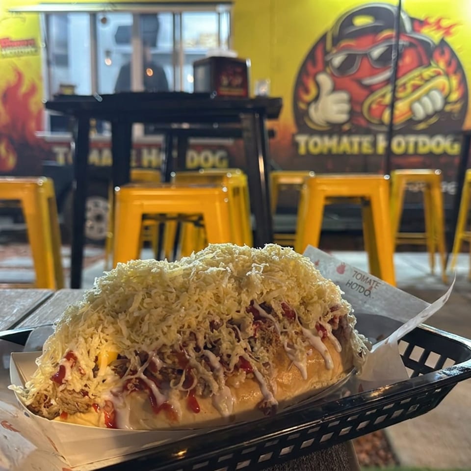 Loaded hot dog topped with shredded cheese and sauces in a basket, with colorful restaurant seating and mural in the background.